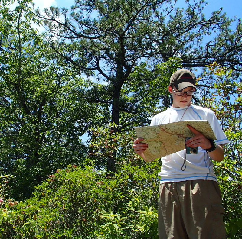 A young man stands in a lush, green forest, engrossed in reading a map. He is wearing a white t-shirt, khaki shorts, glasses, and a cap. A camera hangs around his neck. The surrounding foliage is dense, with various shades of green from the trees and bushes. The sky is visible in the background, peeking through the branches of the trees.